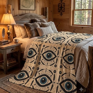 Könnte beinhalten: Eine cremefarbene Wohndecke mit einem sich wiederholenden Muster aus stilisierten blauen und schwarzen Augen, umgeben von strahlenden Linien, kleinen Mondsicheln und federartigen Motiven, drapiert über ein Bett mit strukturierten Kissen und Holzmöbeln.