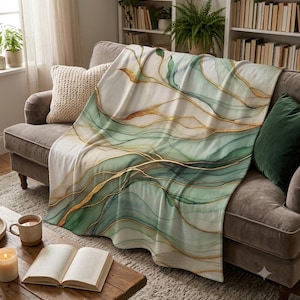 Peut inclure: Un plaid doux présentant un motif abstrait à l'aquarelle avec des couches fluides de vert sauge et de blanc, accentué par de fines lignes dorées scintillantes créant un effet marbré sur la surface du tissu, drapé sur un canapé gris.