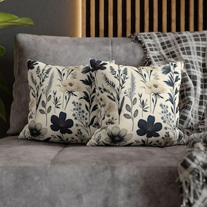 May include: Two square throw pillows with a floral pattern. The pillows feature a cream background with navy blue and light beige botanical designs, including flowers and foliage. The pillows are placed on a gray sofa.