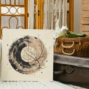 Op de afbeelding: Vierkant canvas kunstwerk met een abstract cirkelvormig ontwerp in zwart en beige tinten, met een takachtig element. Het kunstwerk wordt getoond in een kamer met een rieten mand, een vintage koffer en een bakstenen muur. Thema: interieurdecoratie.