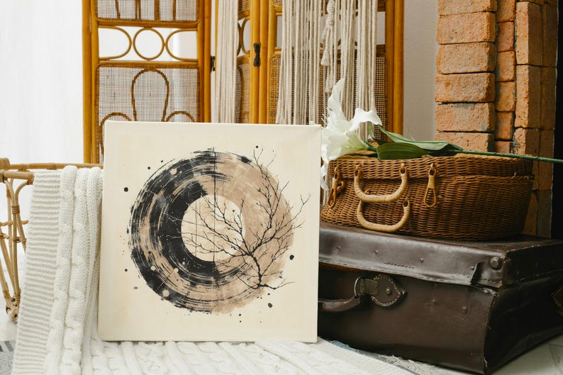 Op de afbeelding: Vierkant canvas kunstwerk met een abstract cirkelvormig ontwerp in zwart en beige tinten, met een takachtig element. Het kunstwerk wordt getoond in een kamer met een rieten mand, een vintage koffer en een bakstenen muur. Thema: interieurdecoratie.