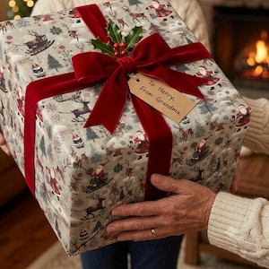 May include: A square gift box wrapped in light gray paper featuring a repeating pattern of Santa Claus, reindeer, and pine trees. The box is tied with a wide, deep red velvet ribbon and topped with a sprig of holly. A tan gift tag reads: To: Henry, From: Grandma.