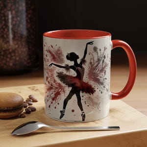 May include: A white ceramic mug with a red rim and handle. The mug features a black silhouette of a ballerina in a red tutu with a watercolor-like background of red, pink, and gray.