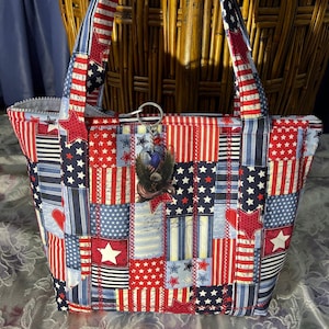 May include: A quilted tote bag featuring a patchwork design of red, white, and blue fabric with stars, stripes, and script patterns. The bag includes two fabric handles and a silver-toned metal keychain depicting an eagle with an American flag.