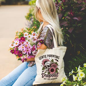 Op de afbeelding: Een witte tote bag met een schedel- en zonnebloemontwerp. De tekst op de tas luidt "Gentle Parenting Dropout Est. This Morning".