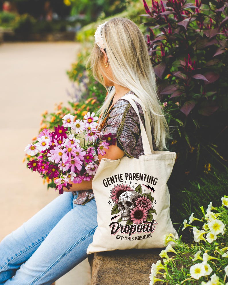 Op de afbeelding: Een witte tote bag met een schedel- en zonnebloemontwerp. De tekst op de tas luidt "Gentle Parenting Dropout Est. This Morning".