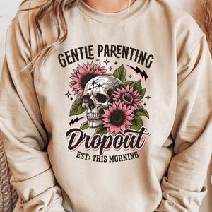 Op de afbeelding: Een beige sweatshirt met de tekst "Gentle Parenting Dropout Est: This Morning" erop gedrukt. De tekst wordt omringd door een schedel en roze bloemen.