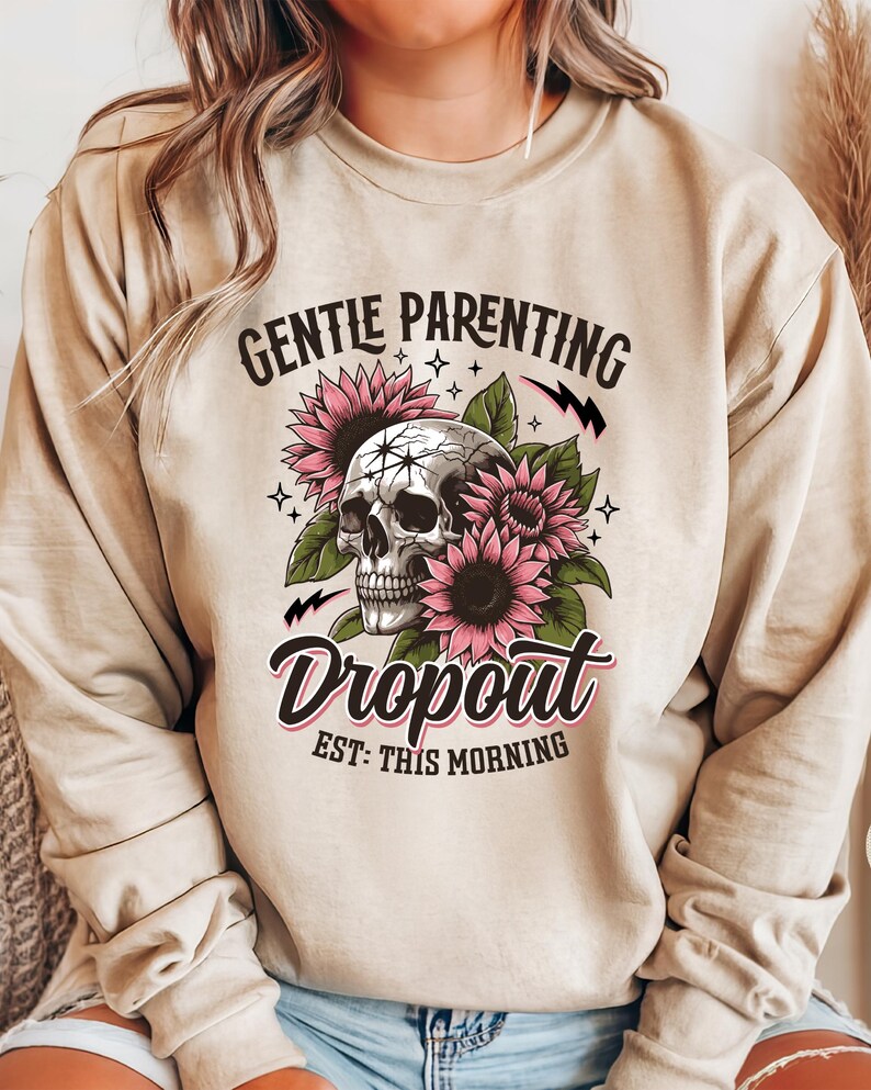 Op de afbeelding: Een beige sweatshirt met de tekst "Gentle Parenting Dropout Est: This Morning" erop gedrukt. De tekst wordt omringd door een schedel en roze bloemen.