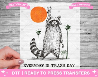 May include: Black and white illustration of a raccoon wearing a peace sign and standing on its hind legs. The raccoon is surrounded by palm trees and a large orange sun. The text "EVERYDAY IS TRASH DAY" is printed below the raccoon.