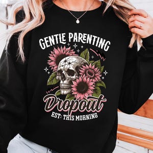 Op de afbeelding: Zwart sweatshirt met een schedel omringd door roze bloemen en de tekst "Gentle Parenting Dropout Est: This Morning".