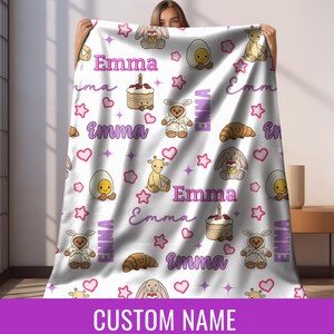 May include: A white blanket with a repeating pattern of cartoon animals, hearts, stars, and the name "Emma" in purple. The blanket features a teddy bear in a bunny suit, a giraffe, a bunny, and a cake. The words "CUSTOM NAME" are at the bottom.