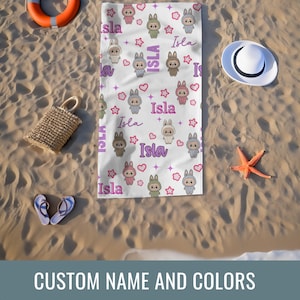 May include: White beach towel featuring a repeating pattern of cartoon bunnies, stars, and hearts. The name "Isla" is written in a script font. The towel is surrounded by beach accessories, including a straw bag, sandals, and a starfish.