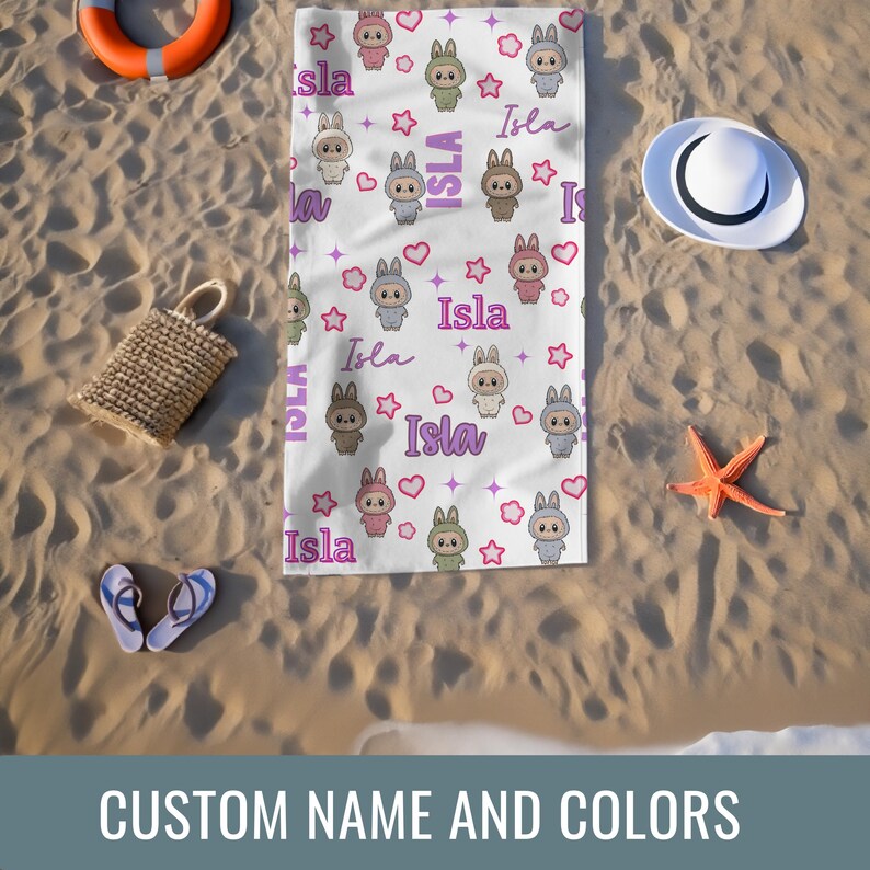 May include: White beach towel featuring a repeating pattern of cartoon bunnies, stars, and hearts. The name "Isla" is written in a script font. The towel is surrounded by beach accessories, including a straw bag, sandals, and a starfish.