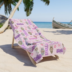 May include: A light pink beach towel with a repeating pattern of cartoon bunnies, stars, and hearts. The word "Stella" is printed in purple throughout the design. The towel is draped over a wooden beach chair on a sandy beach.