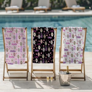 May include: Three personalized beach towels with bunny and star patterns are displayed on wooden beach chairs. The towels feature names like "Stella", "Lilly", and "Isla" in a playful font, with pink, black, and white backgrounds.