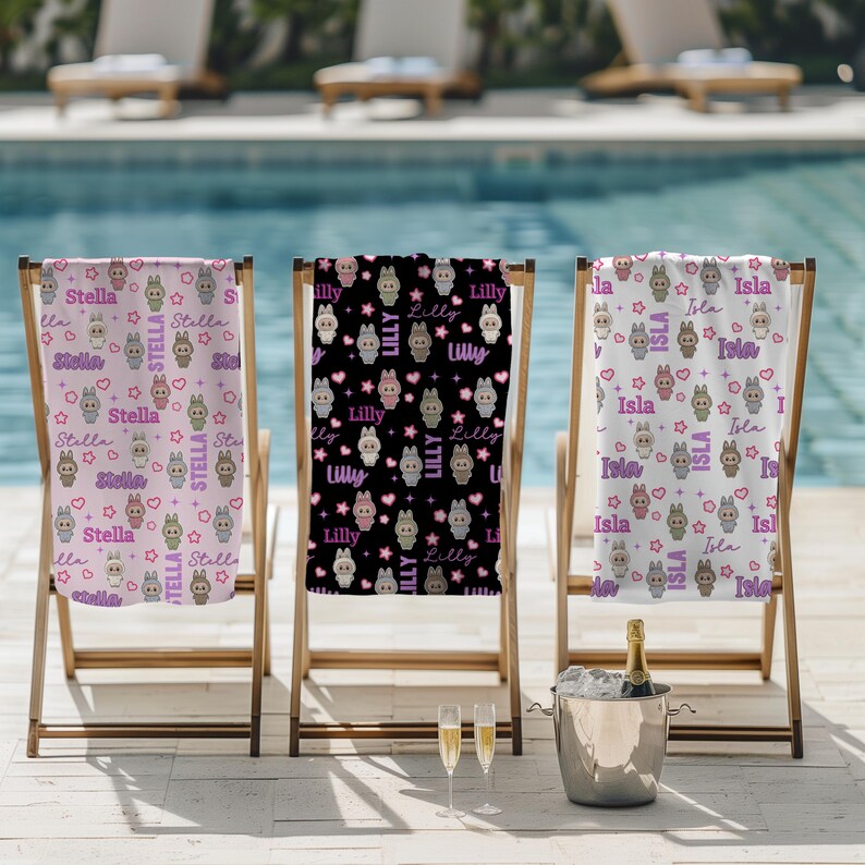May include: Three personalized beach towels with bunny and star patterns are displayed on wooden beach chairs. The towels feature names like "Stella", "Lilly", and "Isla" in a playful font, with pink, black, and white backgrounds.