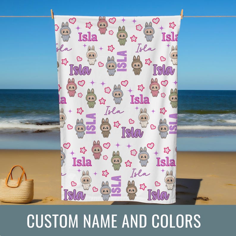 May include: A white beach towel with a repeating pattern of cartoon bunnies, hearts, and stars in various colors. The name "Isla" is written in a script font throughout the design. The towel is hanging on a clothesline on a beach. Text at the bottom says "CUSTOM NAME AND COLORS".