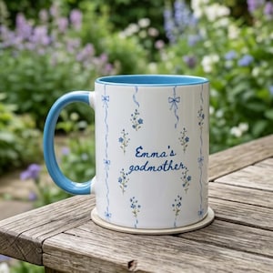 May include: A white ceramic mug with a light blue handle and interior. The exterior features a repeating pattern of vertical blue ribbons with bows and small blue floral sprigs. The text Emma's godmother is printed in blue script on the front.