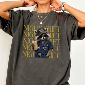 May include: A charcoal grey t-shirt featuring a graphic of a raccoon wearing a black cap with the text NIGHT SHIFT, sunglasses, and a blue medical scrub top with a stethoscope. The background of the graphic repeats the text NIGHT SHIFT in gold.