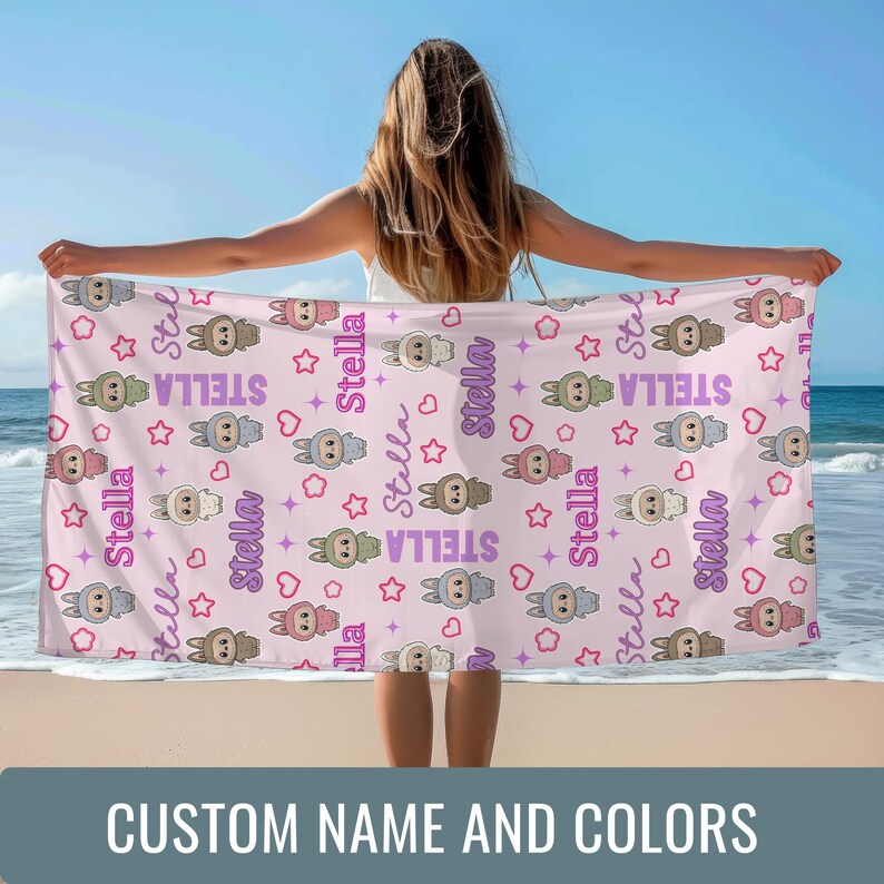 May include: A light pink beach towel with a repeating pattern of cartoon bunnies, stars, and hearts. The name "Stella" is printed in a script font. The towel is held up by a person on a beach. The text "CUSTOM NAME AND COLORS" is at the bottom.
