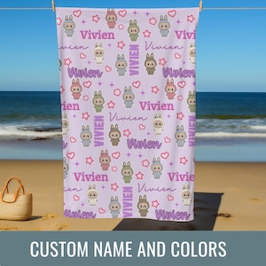 May include: A lavender beach towel with a repeating pattern of cartoon bunnies in various colors, stars, and hearts. The name "Vivien" is written in a cursive font throughout the design. The towel is hanging on a clothesline on a beach.