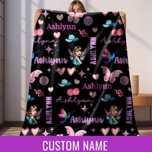 Op de afbeelding: Zwarte deken met een herhalend patroon van harten, vlinders, dobbelstenen, cowboyhoeden, fee&euml;n en de naam "Ashlynn" in roze en wit. De deken heeft de tekst "CUSTOM NAME" onderaan.
