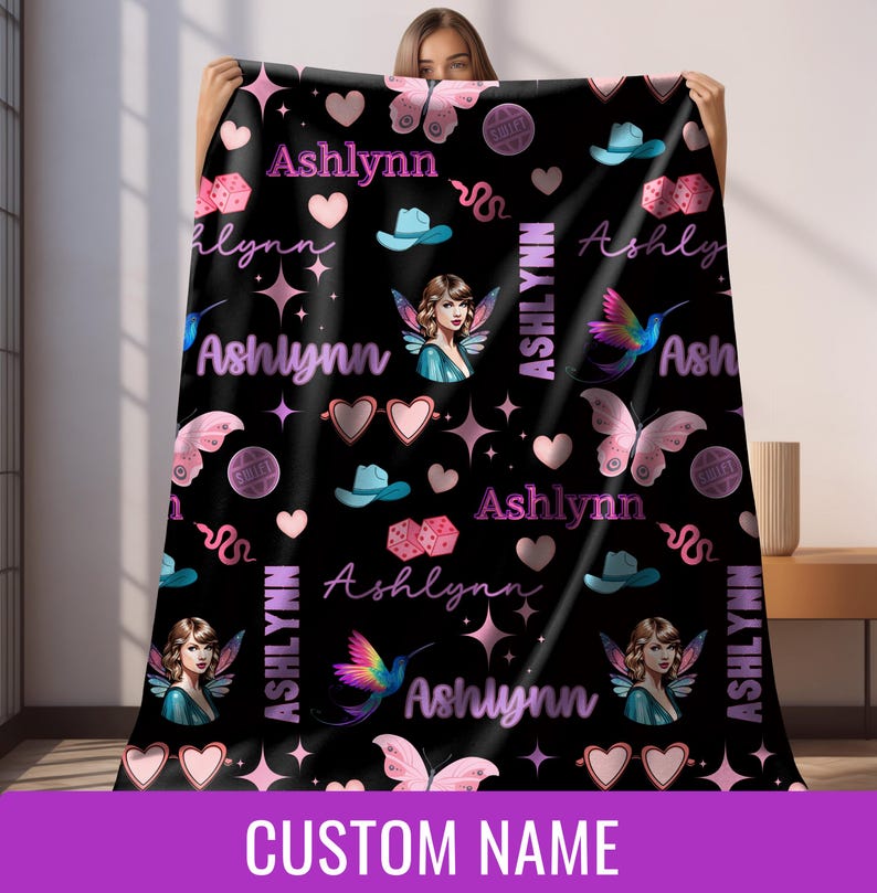 Op de afbeelding: Zwarte deken met een herhalend patroon van harten, vlinders, dobbelstenen, cowboyhoeden, fee&euml;n en de naam "Ashlynn" in roze en wit. De deken heeft de tekst "CUSTOM NAME" onderaan.