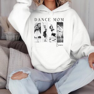 Peut inclure: Un sweat-shirt blanc avec l'inscription "DANCE MOM" au-dessus d'une image en noir et blanc de quatre danseuses. Le nom "Emma" est &eacute;crit en script en dessous de l'image. Le sweat-shirt a une poche avant et une capuche.