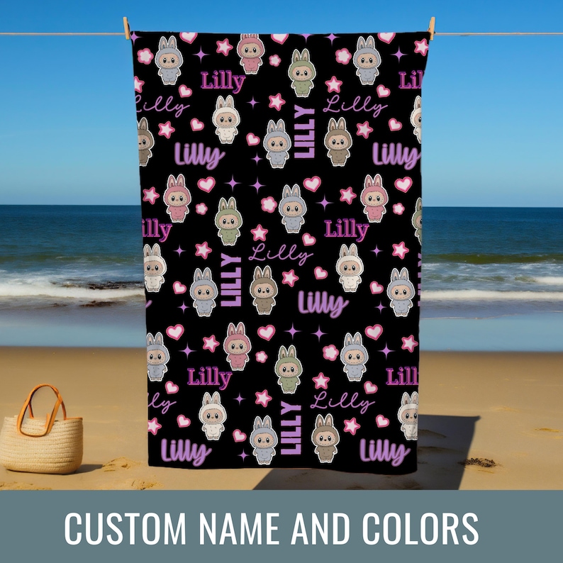 May include: Black beach towel with a repeating pattern of cartoon bunnies in various colors, stars, and hearts. The name "Lilly" is written in a cursive font throughout the design. The towel is hanging on a clothesline on a beach.