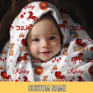 May include: A baby wrapped in a white blanket with a red and gold horse pattern. The blanket has the name "Kirby" and the year "2024" printed on it. The bottom of the image has the text "CUSTOM NAME" in a yellow box.