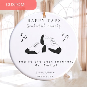May include: White ceramic ornament with a black and white illustration of tap shoes and musical notes. The text reads "HAPPY TAPS Grateful Hearts You're the best teacher, Ms. Emily! From: Emma 2023-2024".