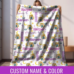 Könnte beinhalten: Weiße Decke mit einem sich wiederholenden Muster aus einer Zeichentrickfigur, Monden, Sternen und dem Namen "Harper" in Lila. Die Decke hat einen lila Rand mit dem Text "CUSTOM NAME & COLOR".