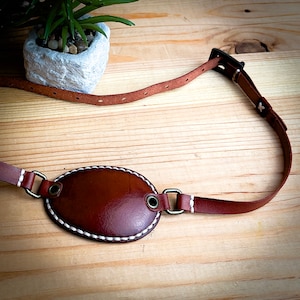May include: A brown leather eye patch with white stitching, attached to a brown leather strap with a metal buckle. The eye patch is oval shaped and has a smooth surface.