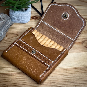 Peut inclure: Étui en cuir marron ouvert avec des coutures blanches et une fermeture à bouton-pression. L'étui contient plusieurs petits objets en bois de couleur claire. L'étui est posé sur une surface en bois.