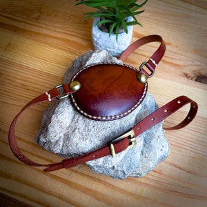 Può includere: Un paraluce per occhio in pelle marrone con un bordo cucito e una fibbia in ottone. Il paraluce per occhio è attaccato a un cinturino in pelle marrone con una fibbia in ottone.