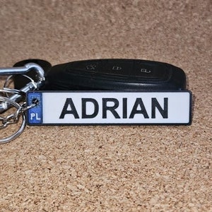 Op de afbeelding: Een sleutelhanger met de naam "ADRIAN" in witte letters gedrukt op een rechthoekig zwart-wit label. De sleutelhanger bevat sleutels, een autosleutel en een zilveren metalen sluiting.