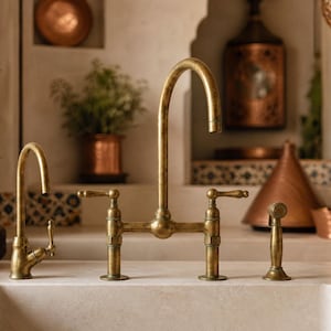 Può includere: Un rubinetto da cucina a ponte con bocca ad arco alto e due maniglie a leva in finitura ottone anticato. Il gruppo rubinetteria include una doccetta laterale separata e un rubinetto secondario più piccolo, montati su un piano in pietra.