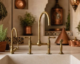 May include: A bridge-style kitchen tap with a high-arc spout and two lever handles in an aged brass finish. The tap assembly includes a separate side sprayer and a smaller secondary tap, all mounted on a light-coloured stone worktop.