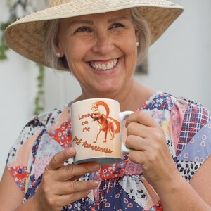 May include: A white ceramic mug with the text "Lean on Me MS Awareness" and a graphic of a dog. The mug is held by a person wearing a straw hat and a colorful patchwork top.
