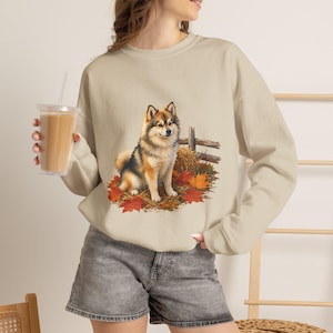 Pode incluir: Uma camisola bege de gola redonda com uma impressão gráfica de um cão fofo castanho e preto sentado num monte de folhas de outono e feno perto de uma cerca de madeira. A camisola tem mangas compridas e gola, punhos e bainha canelados.