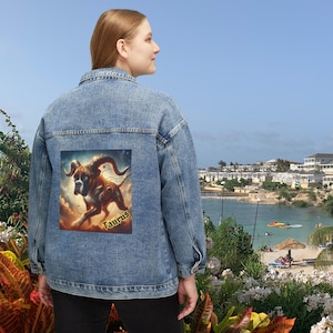 Puede incluir: Chaqueta vaquera azul claro con un gráfico de Tauro colorido en la espalda. El gráfico presenta una criatura parecida a un perro con cuernos y la palabra "Taurus" en dorado. La chaqueta tiene un diseño clásico con botones.