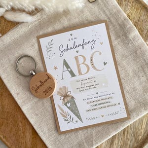 Könnte beinhalten: Ein runder Holzschlüsselanhänger mit der Gravur Schulkind 2026 und eine rechteckige Karte mit dem Text ZUM Schulanfang ABC, Ein neues Kapitel beginnt, Mit Mut, Neugier und ganz viel Freude, Für deinen Weg wünschen wir dir GLÜCKLICHE MOMENTE, LIEBE MENSCHEN UND VIELE KLEINE ERFOLGE.