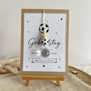 Könnte beinhalten: Eine rechteckige Grußkarte mit Kraftpapier-Rahmen, verziert mit einem Fußball-Motiv und dem Text "Geburtstag", "ALLES LIEBE FÜR DICH" sowie "HAB EINEN TOLLEN TAG!". Ein hängendes Schmuckelement mit Holz- und Fußballperlen sowie einem Kristallanhänger.