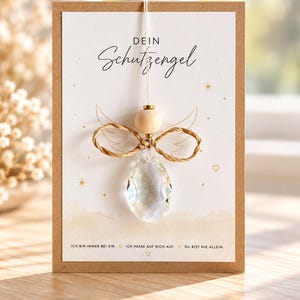 Könnte beinhalten: Eine rechteckige Grußkarte mit Kraftpapierrand, die den Text "DEIN Schutzengel" und ein hängendes Ornament zeigt. Das Ornament besteht aus einer Holzperle, einer Juteschleife und einem klaren, facettierten Kristall in Tropfenform.