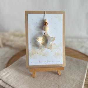 Könnte beinhalten: Ein transparenter, facettierter sternförmiger Anhänger hängt an einer Schnur mit drei Holzperlen auf einer rechteckigen Karte, die mit Wolken und Sternen illustriert ist und den Text "kleiner Stern" unten auf der Karte trägt.