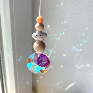 Könnte beinhalten: Ein hängendes Sonnenfänger-Ornament mit einer weißen Schnur, einer kleinen pfirsichfarbenen Perle, einer größeren beigen Perle, einer weißen wolkenförmigen Perle mit lächelndem Gesicht, einer weiteren beigen Perle und einem runden, facettierten Prisma.