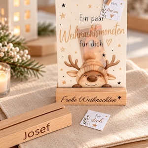 Könnte beinhalten: Eine helle Karte mit Rentier-Illustration, dem Text 'Ein paar Weihnachtsmoneten für dich' und einem Anhänger mit 'Von Herzen'. Sie steht in einem Holzsockel mit der Gravur 'Frohe Weihnachten'. Ein separater Holzblock ist mit 'Josef' graviert.