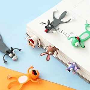 May include: A collection of whimsical animal-shaped bookmarks, including a gray wolf, a white dog, a brown rabbit, a green frog, a purple mouse, a black donkey, and an orange creature. Each bookmark is designed to peek out from the pages of a book, adding a playful touch.