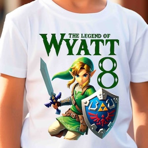 Puede incluir: Camiseta blanca con un gráfico verde de un personaje de videojuego con espada y escudo. El texto "THE LEGEND OF WYATT 8" está encima del personaje. La parte trasera de la camiseta tiene un diseño verde con las palabras "BIRTHDAY BOY". 100% algodón.
