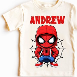 May include: A white vintage t-shirt with a cartoon image of Spiderman wearing a red and blue hoodie. The text "ANDREW" is printed in red above the image.